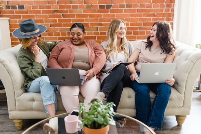 Four women sitting on the couch and laughing while working on their laptops.